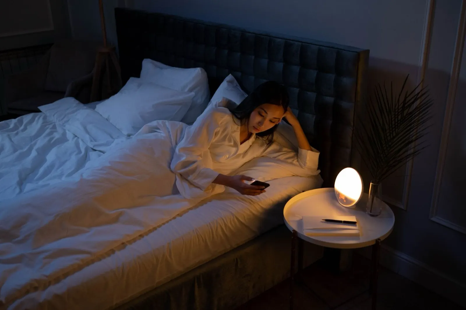 A woman awake late at night in a dark bedroom, lit only by a bedside lamp and her phone screen
