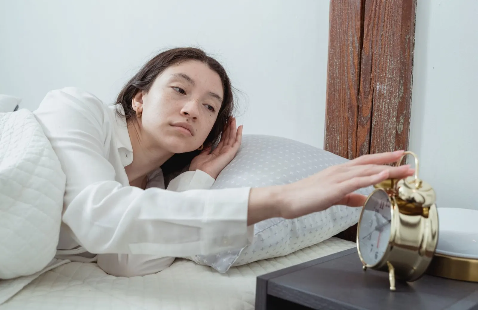 A tired woman lying in bed reaching out to switch off an alarm clock on the bedside table