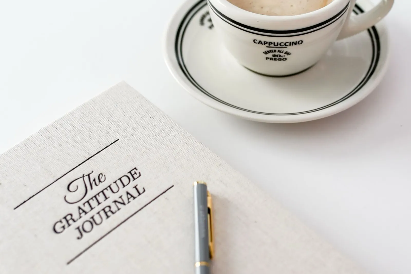 A gratitude journal, a pen and a cup of coffee arranged on a white table